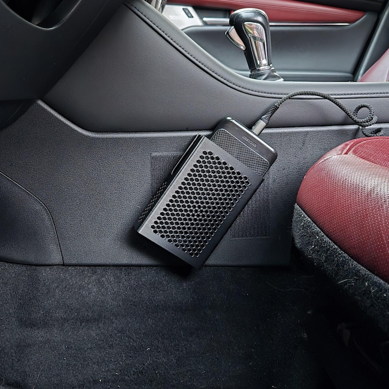 May include: A black rectangular electronic device with a honeycomb-patterned front grille, mounted to the side of a car center console. The device is connected by a black coiled cable and features a textured top surface with a small logo.