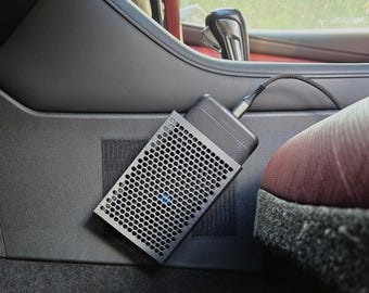 May include: A black electronic device with a perforated metal cover is plugged into a car's interior. The device is connected to a cable. The car's interior is black and gray, with a red seat visible.