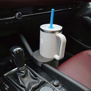 May include: A white insulated mug with a handle and a blue straw, in a car cup holder. The mug has a silver lid and a white logo. The car interior features a red seat and black dashboard.
