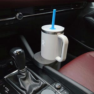 May include: A white insulated mug with a handle and a blue straw, placed in a car's cup holder. The mug has a silver rim and a white lid. The car interior features a red seat and a black gear shift.