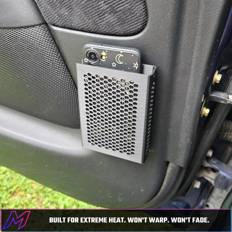 May include: A black rectangular car door storage pocket with a honeycomb mesh front panel, holding a smartphone with a celestial-themed case. The text at the bottom reads: BUILT FOR EXTREME HEAT. WON'T WARP. WON'T FADE.