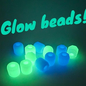 May include: A collection of glow-in-the-dark beads in shades of blue and green. The words "Glow beads!" are written in bright green above the beads. The beads are cylindrical with a hole through the centre, and glow brightly.