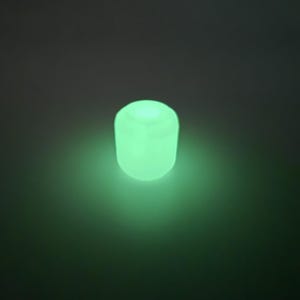 May include: A glowing, cylindrical object emits a soft green light against a dark background. The object appears to be made of a translucent material, with a rounded top and a slightly textured surface. The overall effect is one of gentle illumination.