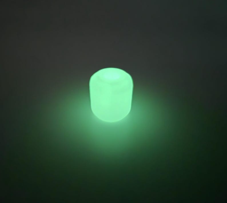 May include: A glowing, cylindrical object emits a soft green light against a dark background. The object appears to be made of a translucent material, with a rounded top and a slightly textured surface. The overall effect is one of gentle illumination.
