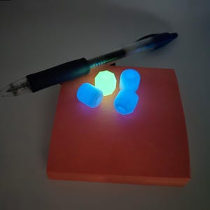 May include: A clear pen rests on an orange sticky note pad. Three glowing blue beads and one glowing green bead are on the pad. The beads and the pad are illuminated in a dark setting.