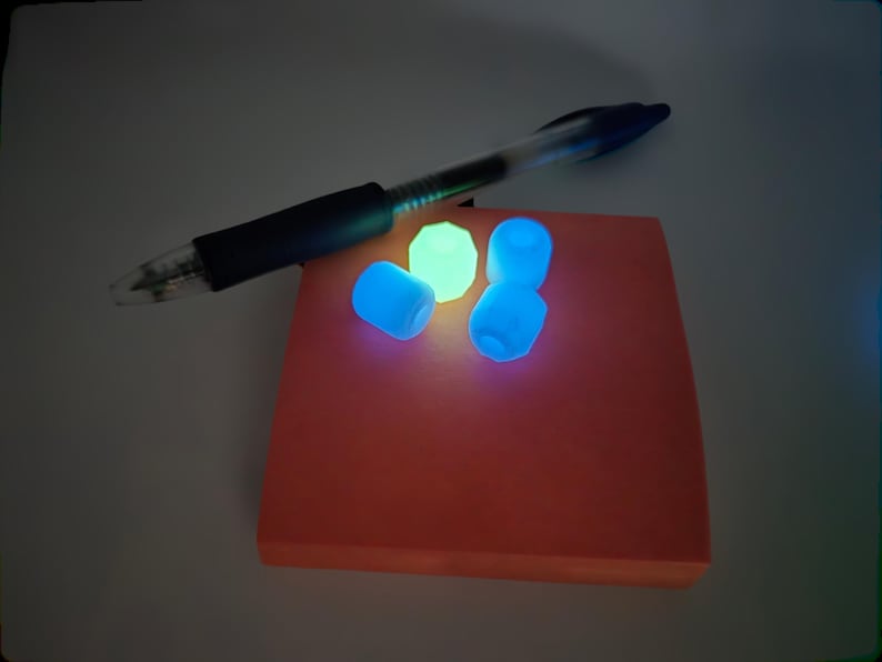 May include: A clear pen rests on an orange sticky note pad. Three glowing blue beads and one glowing green bead are on the pad. The beads and the pad are illuminated in a dark setting.