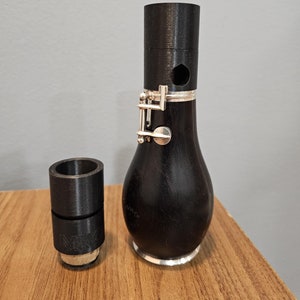 Pode incluir: Um oboé de madeira preta com uma chave de prata e uma boquilha de plástico preto. O oboé está sobre uma superfície de madeira.