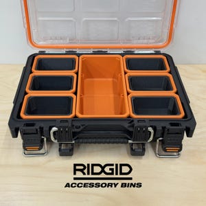 May include: An open black and orange RIDGID accessory bin with a clear lid. The bin contains several black and orange storage compartments. The text "RIDGID ACCESSORY BINS" is printed in white on the bottom of the image.