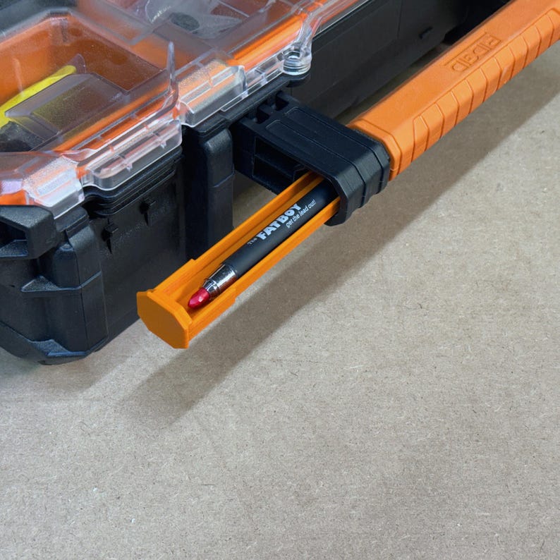 Peut inclure: Organisateur d'outils noir et orange avec un couvercle transparent. Un stylo noir avec le texte "FATBOY per the read out" est dans un support orange. La poign&eacute;e de l'organisateur d'outils est orange et noire.