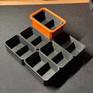 May include: An assortment of black and orange storage containers. The containers are rectangular with internal dividers, creating multiple compartments. One container is orange, while the others are black. The containers are designed for organizing small items.
