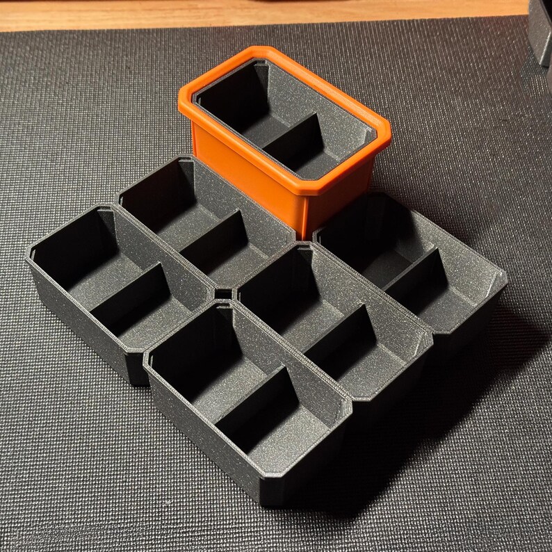 May include: An assortment of black and orange storage containers. The containers are rectangular with internal dividers, creating multiple compartments. One container is orange, while the others are black. The containers are designed for organizing small items.