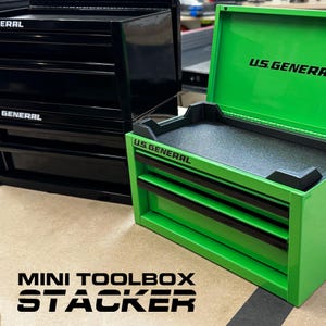 Könnte beinhalten: Ein hellgrüner U.S. General Mini-Werkzeugkasten mit Klappdeckel, schwarzen Schubladengriffen und einer strukturierten schwarzen Innenschale, neben einem passenden schwarzen U.S. General Mini-Werkzeugkasten mit aufgedrucktem Markennamen.