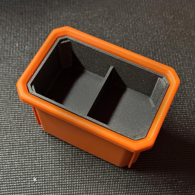 May include: An orange and black rectangular container with two compartments. The container has a black interior and a raised orange border. The item appears to be 3D printed.