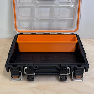 May include: An open storage case featuring a black base, a clear lid with an orange seal, and a removable orange rectangular tray insert. The case includes heavy-duty metal latches on the front and a sturdy handle for secure transport and organization.