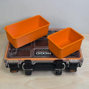 May include: A black and clear Ridgid Pro Gear System Gen 2.0 tool case with two rectangular orange plastic storage bins placed on top. The tool case features a transparent lid, black latches, and a central handle with the brand name printed in white.