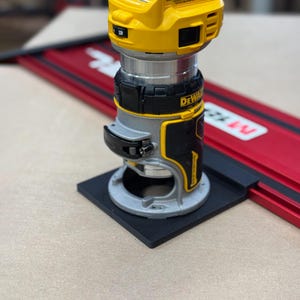 Puede incluir: Una fresadora DeWalt amarilla y negra con base plateada, montada sobre una base cuadrada negra. La fresadora está colocada sobre un sistema de rieles rojo y negro. La imagen está tomada desde un ángulo de cerca.