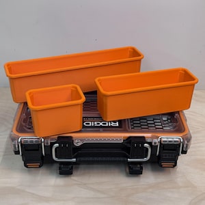 以下が含まれることがあります： 黒と透明のRidgid製ツールボックスと、その上に置かれた3つのオレンジ色のプラスチック製収納ケース。ツールボックスには金属製のハンドル、黒いラッチ、透明な蓋が付いています。オレンジ色のケースは長方形と正方形で、整理整頓用に設計されています。
