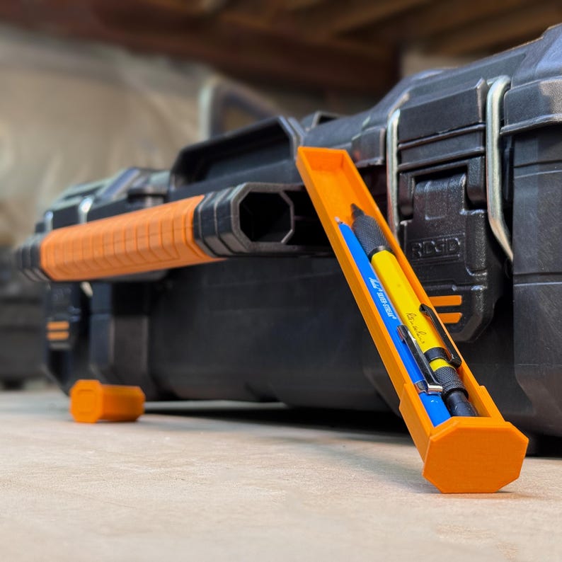 Peut inclure: Mallette &agrave; outils noire et orange avec un porte-crayons orange. Le porte-crayons contient un crayon bleu et un crayon jaune. La mallette a une poign&eacute;e orange et la marque "RIDGID" est visible sur le c&ocirc;t&eacute;.