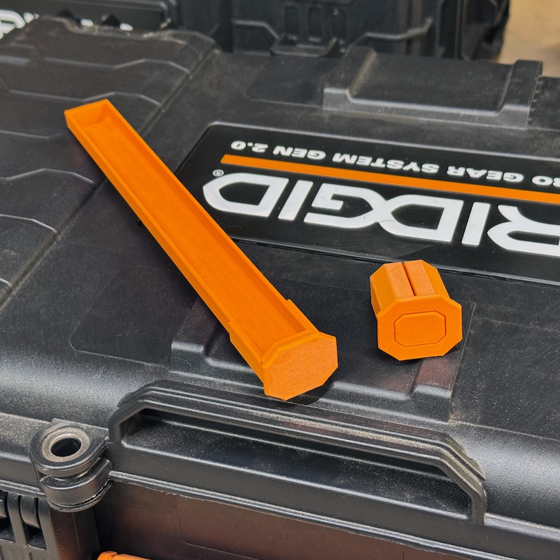 Peut inclure: Deux accessoires en plastique orange, l'un long et rectangulaire, l'autre de forme hexagonale, sont pos&eacute;s sur une bo&icirc;te &agrave; outils noire. La bo&icirc;te &agrave; outils porte le nom de la marque "RIDGID" en blanc.