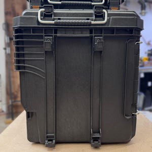 May include: Black, heavy-duty tool storage case with a textured surface. Features a top handle, metal latches, and side clips. The case has a rectangular shape with a ridged design on the side. Another similar case is stacked on top.