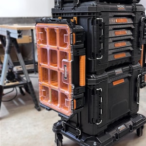 Può includere: Un sistema modulare di stoccaggio attrezzi Ridgid nero su ruote, dotato di un organizer verticale con coperchio arancione trasparente e scomparti multipli fissati al lato di una pila di robuste valigette nere con dettagli arancioni.