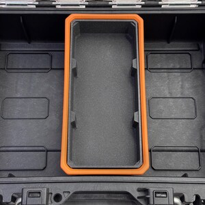 May include: Black storage case with an orange-trimmed compartment. The interior features a textured, dark gray surface. Rectangular compartments are visible within the case, suggesting organized storage. The case is designed for protection and organization.