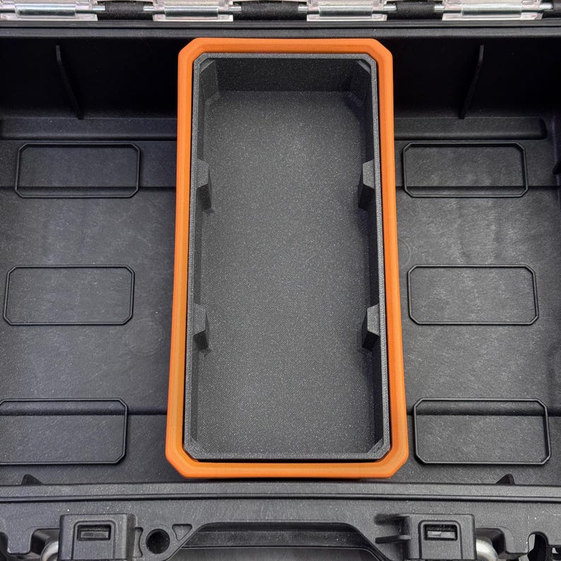 May include: Black storage case with an orange-trimmed compartment. The interior features a textured, dark gray surface. Rectangular compartments are visible within the case, suggesting organized storage. The case is designed for protection and organization.