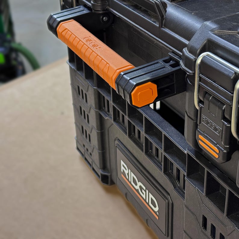 Peut inclure: Gros plan sur une bo&icirc;te &agrave; outils noire RIDGID avec une poign&eacute;e orange. La poign&eacute;e est textur&eacute;e pour une meilleure prise et porte le logo RIDGID. La bo&icirc;te a une conception robuste avec plusieurs compartiments et des loquets s&eacute;curis&eacute;s.