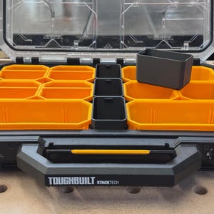 Puede incluir: Un maletín organizador de herramientas ToughBuilt StackTech negro con tapa transparente, que incluye múltiples compartimentos extraíbles amarillos y negros. El maletín tiene un asa negra resistente con un detalle amarillo y el nombre de la marca.
