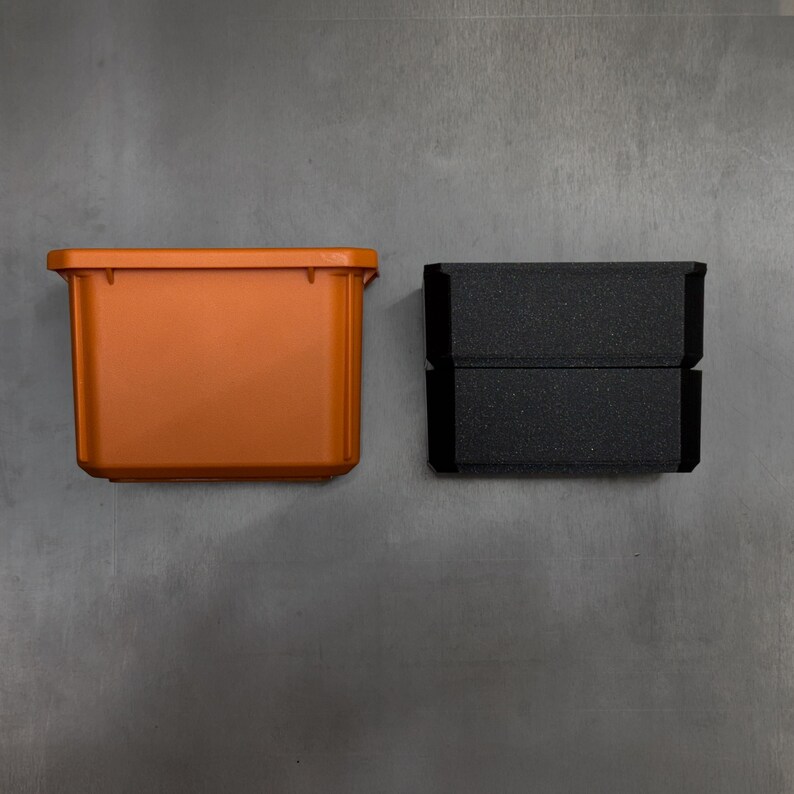 May include: An orange plastic container and a black foam insert. The container has a rectangular shape and a lip around the top edge. The foam insert is made of two stacked pieces. The container is likely used for storage.
