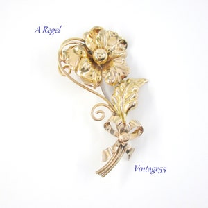 May include: A gold-toned floral brooch with a flower, leaves, and a bow. The brooch has a detailed design with a polished finish. The text "A Regel" and "Vintage55" are visible on the image.