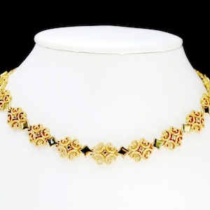 May include: Gold-toned necklace with ornate, symmetrical floral designs and red and black accents. The necklace is displayed on a white mannequin bust against a black background. Jewelry.