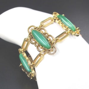 May include: A gold-tone link bracelet featuring three oval-shaped green cabochon stones. Each stone is set within an ornate, filigree-style metal frame, connected by smooth, rectangular gold-tone metal links that form the chain of the bracelet.