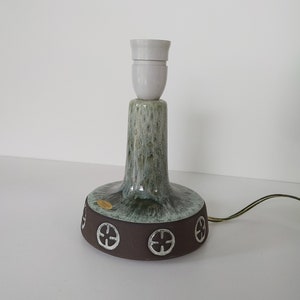 Puede incluir: Una lámpara de mesa de cerámica verde y marrón con una pantalla de lámpara de cerámica blanca. La base de la lámpara tiene un diseño circular con cuatro círculos.