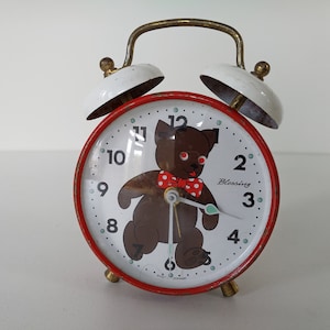 Könnte beinhalten: Roter und weißer Wecker mit Teddybär-Motiv. Das Zifferblatt hat schwarze Zahlen und einen braunen Teddybär mit einer rot-weiß gepunkteten Schleife. Das Zifferblatt trägt außerdem die Aufschrift "Blessing".