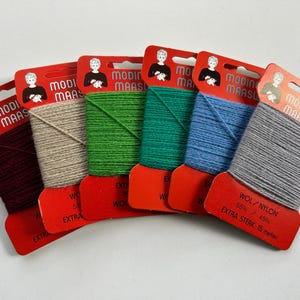 Puede incluir: Una colección de ovillos de hilo de colores sobre etiquetas de cartón rojas y naranjas. Los ovillos son en tonos burdeos, beige, verde, turquesa, azul y gris. Las etiquetas tienen el texto "MODI MAAS" y "WOL/NYLON EXTRA STERK 15 meter".