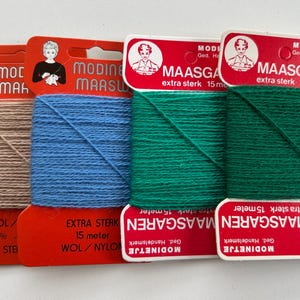 Op de afbeelding: Vier spoelen garen in verschillende kleuren: bruin, blauw en twee groentinten. Elke spoel heeft een label met de tekst "MAASGAREN" en een vintage illustratie van een vrouw. De labels vermelden ook "EXTRA STERK".