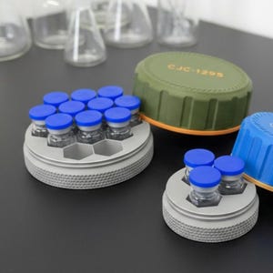 May include: Two gray circular vial holders with hexagonal slots containing small glass vials with blue caps. One holder is paired with a large olive green lid labeled CJC-1295, and the other with a blue lid labeled GLP-1, both featuring textured edges.