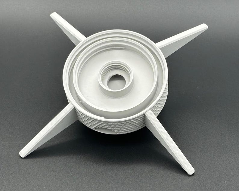 May include: A light gray circular component featuring a central threaded opening and four radiating pointed arms. The outer edge of the cylindrical base displays a textured, diamond-patterned grip, providing a functional design for this mechanical part.