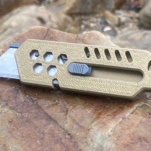 May include: A tan utility knife with a silver blade and a grey slider. The knife has a textured surface and a series of holes and slots. The blade is partially extended, ready for use.