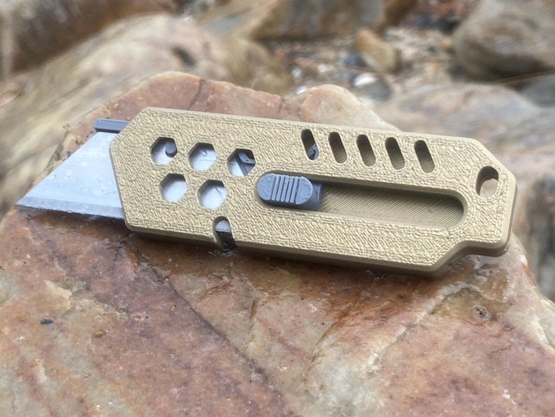 May include: A tan utility knife with a silver blade and a grey slider. The knife has a textured surface and a series of holes and slots. The blade is partially extended, ready for use.