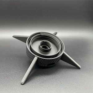 A black, 3D-printed object with a central circular design and four radiating arms. The central portion has concentric rings and a textured surface. The arms are flat and extend outward, creating a star-like appearance.