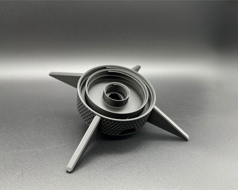 A black, 3D-printed object with a central circular design and four radiating arms. The central portion has concentric rings and a textured surface. The arms are flat and extend outward, creating a star-like appearance.