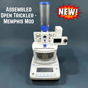 May include: An assembled Open Trickler Memphis Mod powder dispenser featuring a clear vertical hopper with a blue base, a white housing, and a digital control screen labeled OpenTrickler. The unit sits atop a white A&D precision scale base.