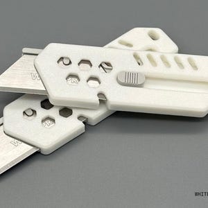close up of two stylized, white utility knives with the blade extended in a ready-to-use position laying against a white background. The knife has a gray button for the sliding mechanism and decorative holes and diagonal slats on the top.