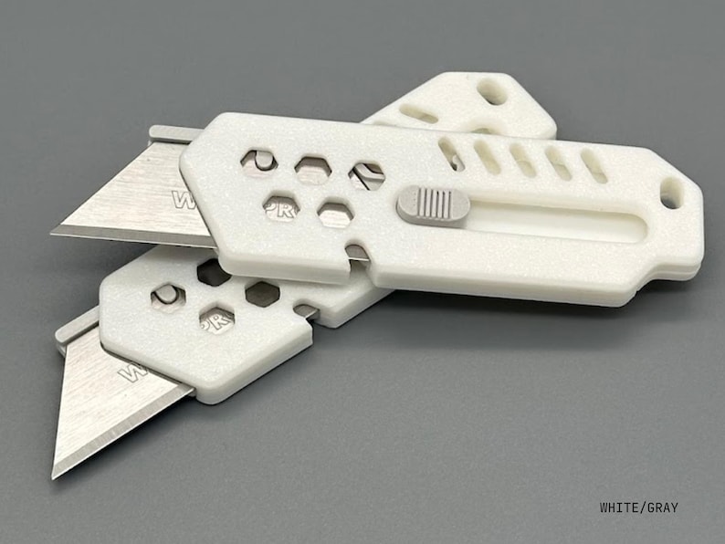 close up of two stylized, white utility knives with the blade extended in a ready-to-use position laying against a white background. The knife has a gray button for the sliding mechanism and decorative holes and diagonal slats on the top.