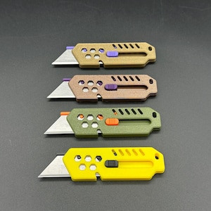 four, stylized, colored utility knives lay against a gray background. their blades are extended in a ready-to-use position and each knife has decorative holes and diagonal slats on the top. The knives have different colored buttons for the slide mechanism of the blade. The colors are (top-bottom): bronze, copper, dark green, and yellow.