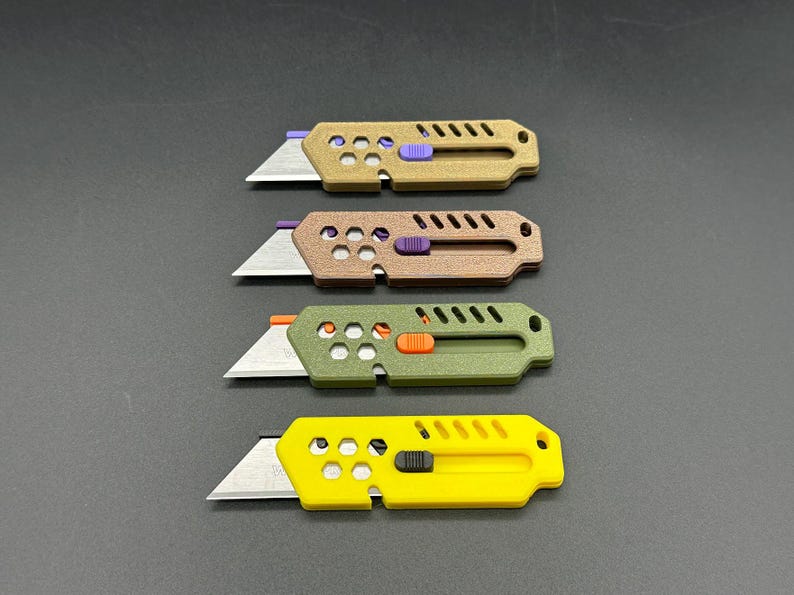 four, stylized, colored utility knives lay against a gray background. their blades are extended in a ready-to-use position and each knife has decorative holes and diagonal slats on the top. The knives have different colored buttons for the slide mechanism of the blade. The colors are (top-bottom): bronze, copper, dark green, and yellow.