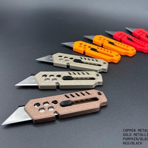 seven, colored, stylized utility knives laid out in the shape of a wave against a gray background. the blades are extended on all in a ready-to-use position. the colors of the knives are:  1 copper, 2 gold, 2 pumpkin, and 2 red.  Each knife has decorative holes and slats.
