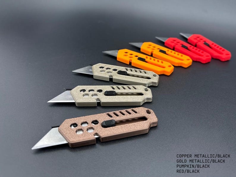 seven, colored, stylized utility knives laid out in the shape of a wave against a gray background. the blades are extended on all in a ready-to-use position. the colors of the knives are:  1 copper, 2 gold, 2 pumpkin, and 2 red.  Each knife has decorative holes and slats.